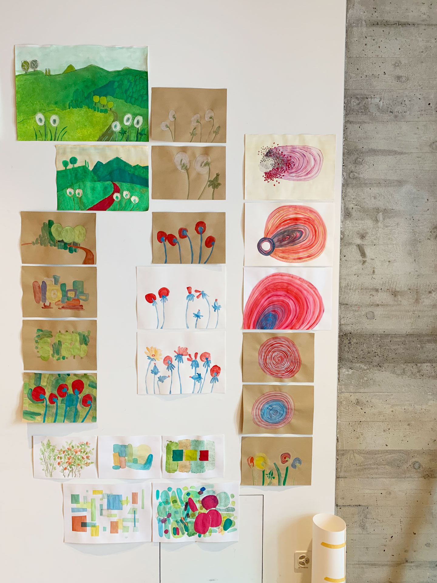 Wandansicht im Atelier: Mehrere Reihen von Zeichnungen und Aquarellen mit Landschaften, abstrakten Blumen und farbigen Formen sind links auf einer weissen Wand arrangiert, rechts schliesst eine rohe Betonwand an.
