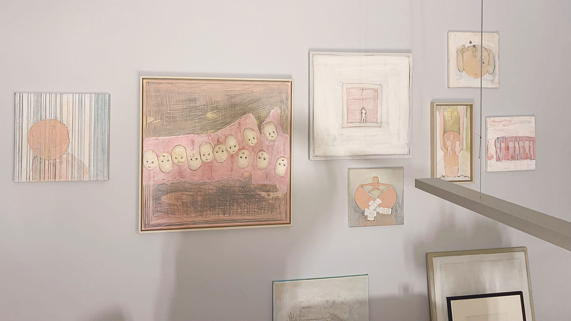 Wandinstallation mit mehreren gerahmten Zeichnungen und Malereien in Rosa- und Beigetönen in einem Ausstellungsraum, darunter ein grosses Bild mit Köpfen in einer Reihe und kleinere figurative Motive.