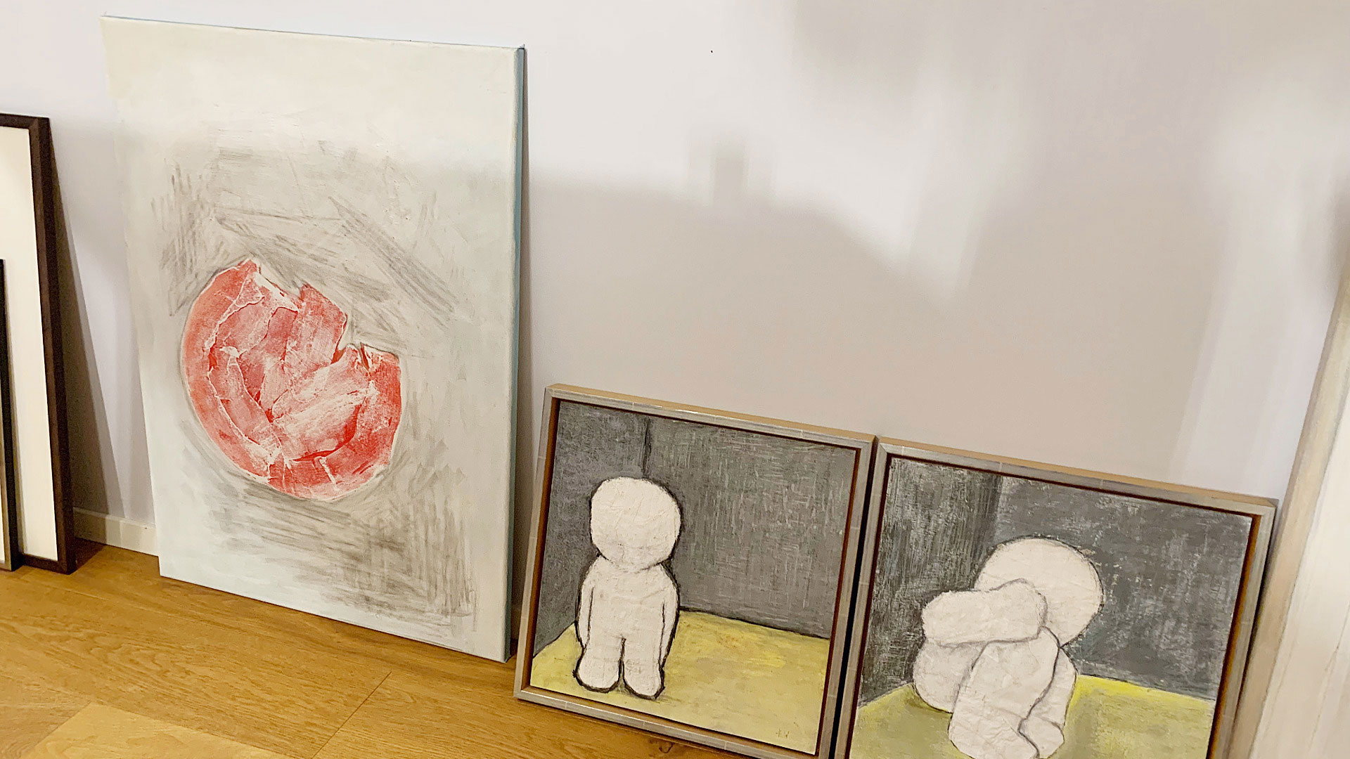 Drei an die Wand gelehnte Bilder auf Parkettboden: links ein grosses weisses Hochformat mit einer rot-fragmentierten Kreisform auf rauem Grund, daneben zwei kleinere, gerahmte Quadrate mit weissen, Figuren vor grauem Hintergrund und gelbem Boden.​