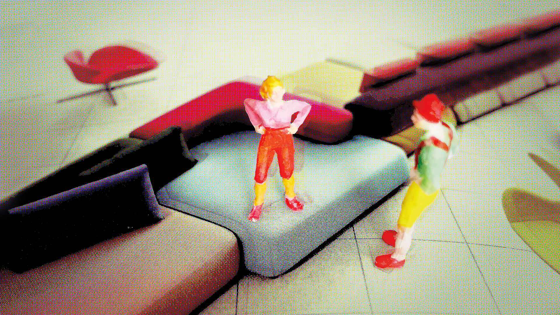 Zwei bunte Modellfiguren stehen auf gepolsterten Sitzmodulen in kräftigen Farben; im Hintergrund sind weitere Möbel und ein roter Lounge-Sessel zu sehen.