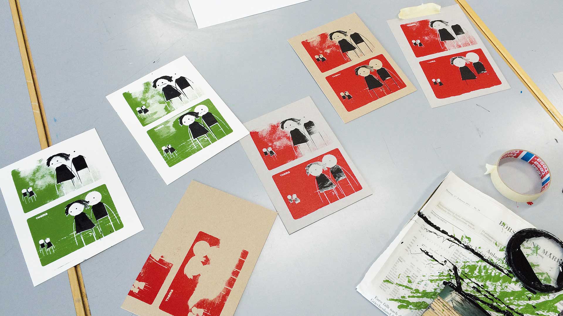 Mehrere im Siebdruck entstandene Kairos-Kartenbögen mit roten und grünen Hintergrundflächen und Figurenpaaren liegen zum Trocknen auf dem Arbeitstisch, umgeben von Klebebandrolle und Farbspritzern.