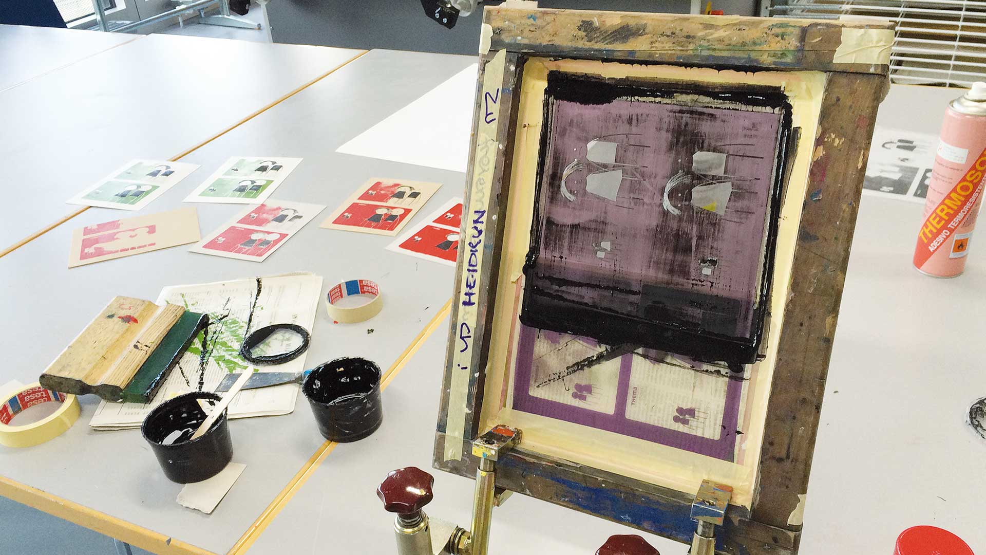 Siebdruckrahmen mit violetter Druckschablone für Kairos-Karten auf einem Arbeitstisch, umgeben von Farbbechern, Rakel und ausgedruckten Kartenbögen, die den analogen Druckprozess der Illustrationen zeigen.