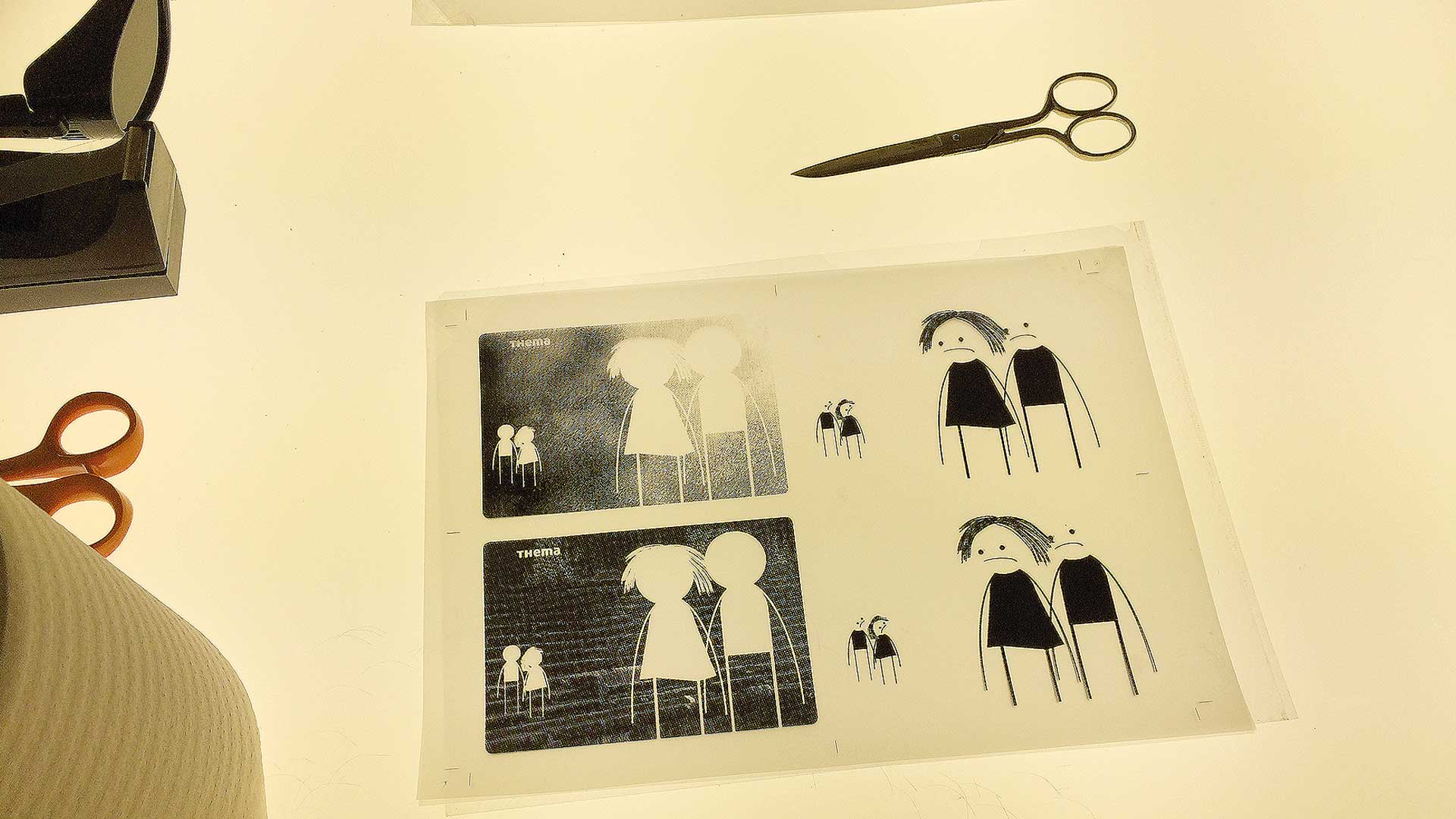 Transparentfolie mit Kairos-Kartenmotiven und freigestellten Figurenpaaren auf einem Leuchttisch, daneben Schere und Arbeitsmaterialien als Teil der Siebdruckvorbereitung.
