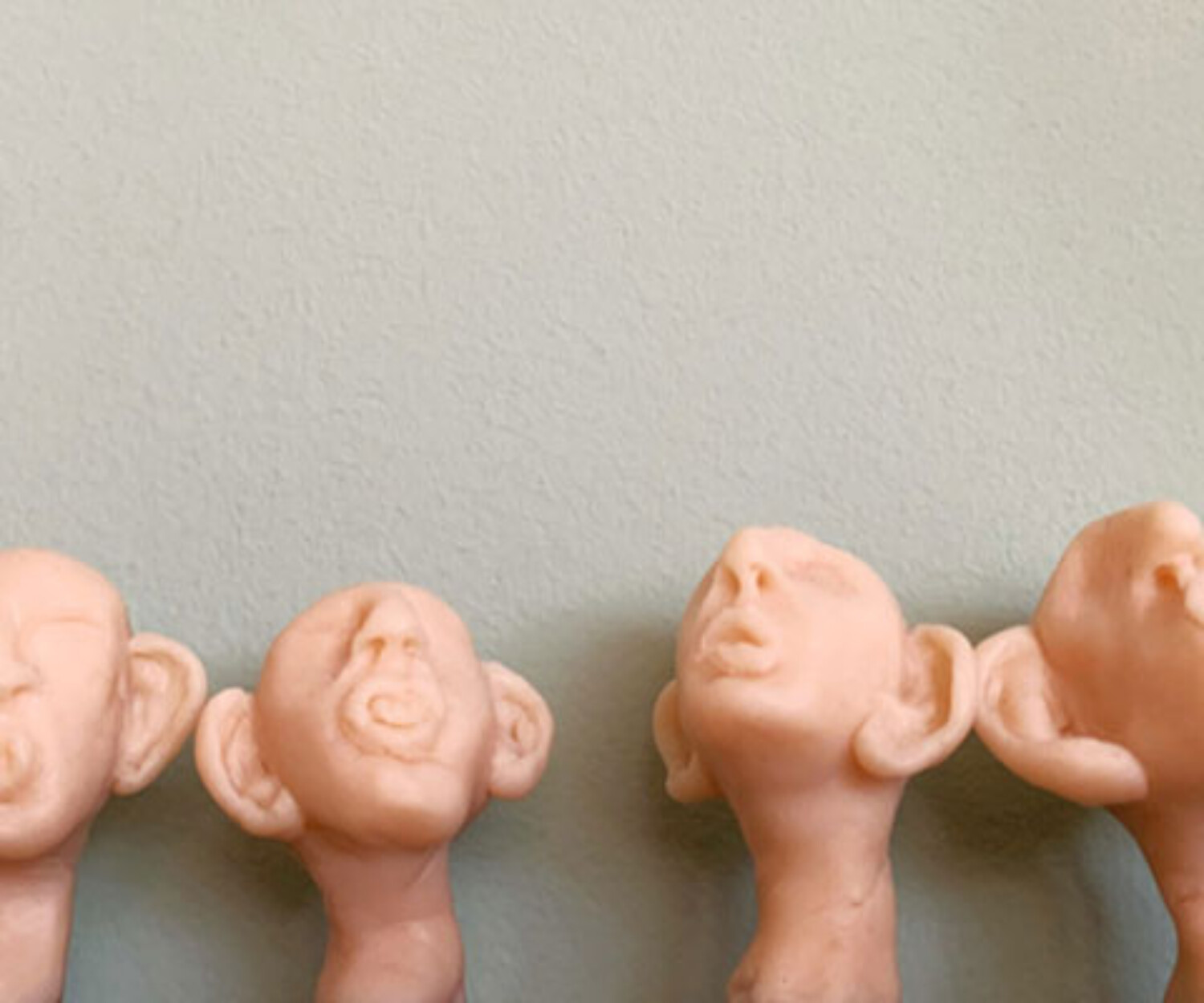 Vier hautfarbene Kopfskulpturen mit übergrossen Ohren blicken nach oben und lauschen, vor einer hellen Wand.