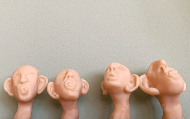 Vier hautfarbene Kopfskulpturen mit übergrossen Ohren blicken nach oben und lauschen, vor einer hellen Wand.