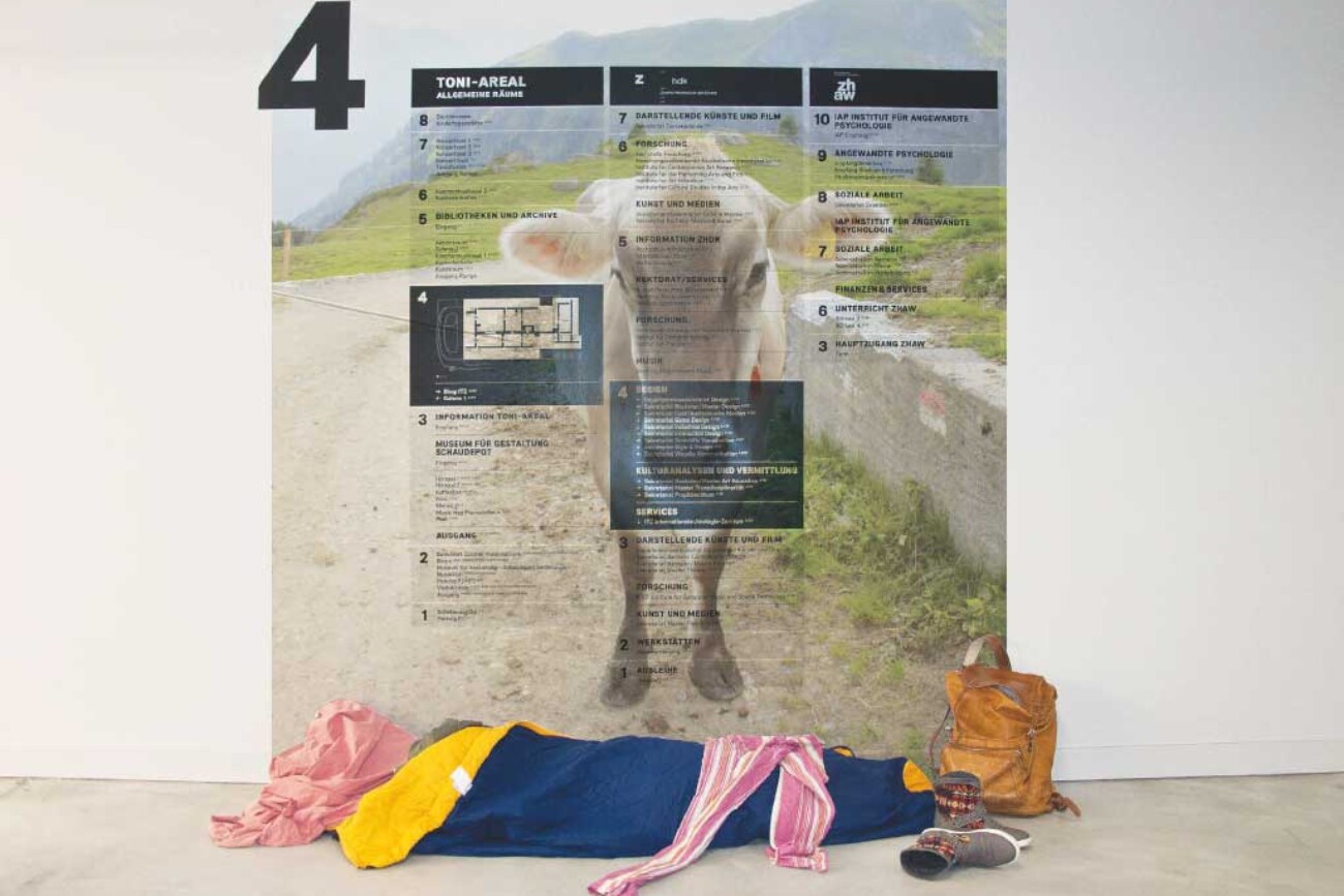 Person im Schlafsack liegt vor einer grossformatigen Wandinstallation im Toni-Areal, die einen eine Kuh und einen Gebäudeübersichtsplan kombiniert und den nüchternen Gang in eine träumerische Landschaft verwandelt.