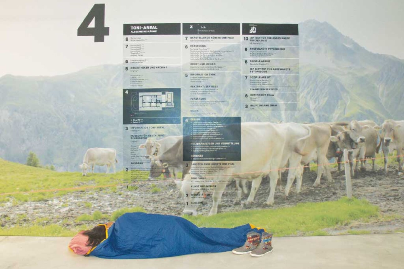 Person im Schlafsack liegt vor einer grossformatigen Wandinstallation im Toni-Areal, die einen Alpwiesenplan mit Kühen und Gebäudeübersichtsplan kombiniert und den nüchternen Gang in eine träumerische Landschaft verwandelt.
