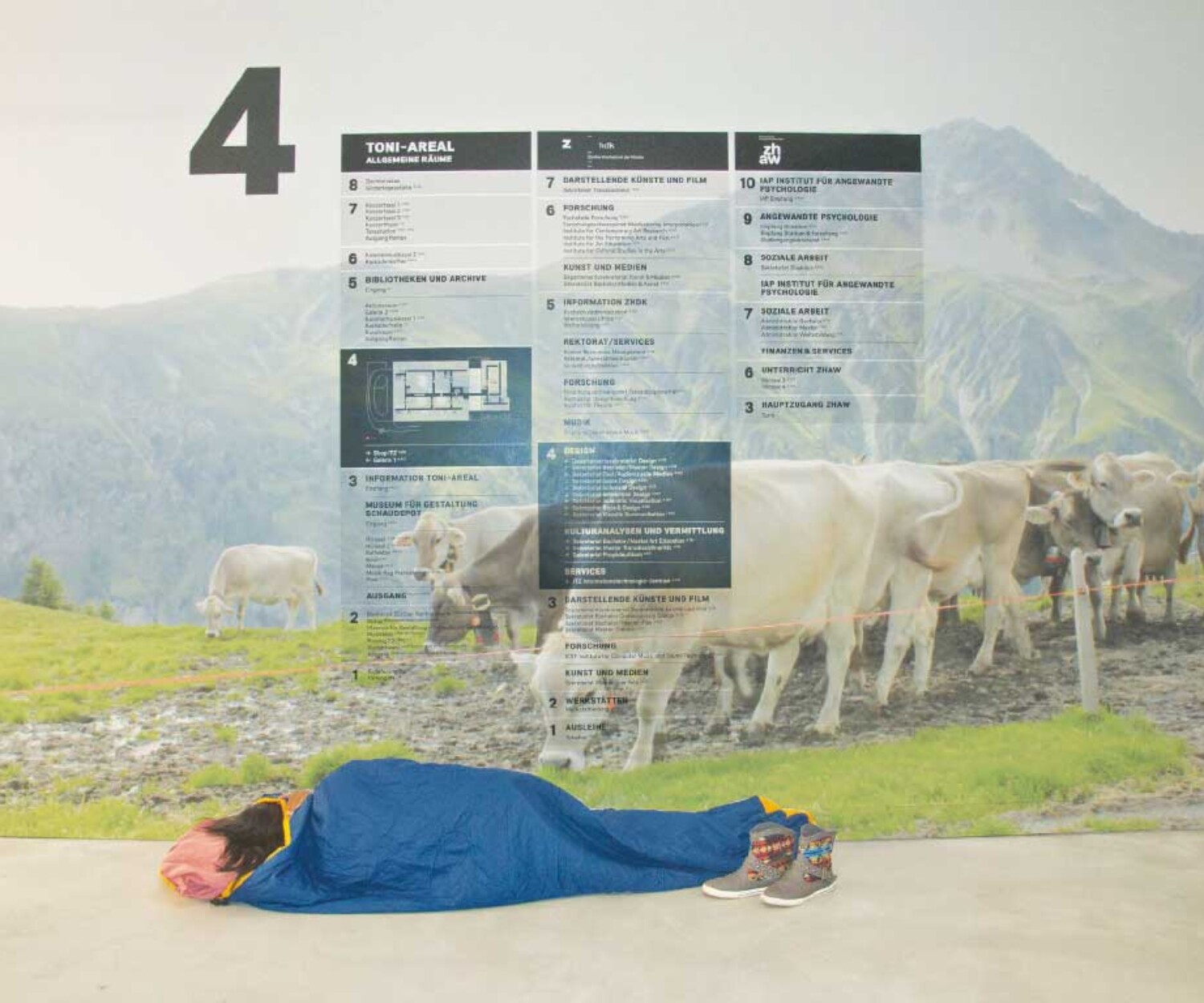 Person im Schlafsack liegt vor einer grossformatigen Wandinstallation im Toni-Areal, die einen Alpwiesenplan mit Kühen und Gebäudeübersichtsplan kombiniert und den nüchternen Gang in eine träumerische Landschaft verwandelt.