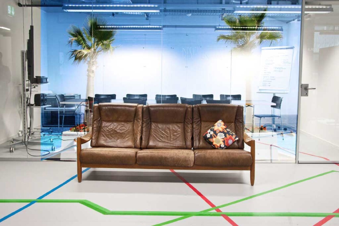 Braunes Ledersofa im Atelier des Toni-Areals, dahinter ein verglaster Sitzungsraum mit Palmen, Strandprojektion und blauer Lichtstimmung.
