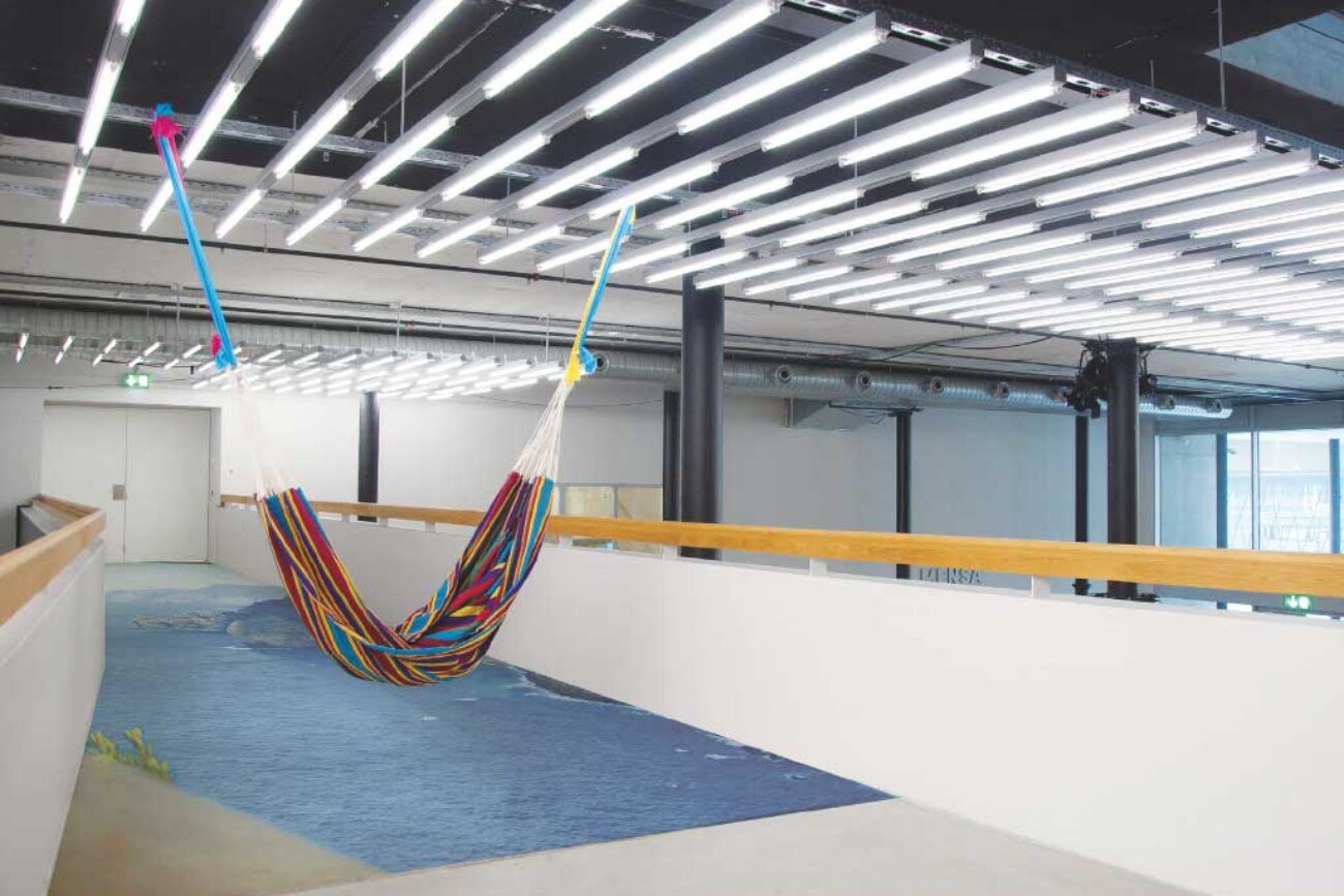 Farbige Hängematte auf der Passerelle im Toni-Areal, schräg in den Raum gespannt, darunter eine blaue Bodeninstallation, die den grauen Korridor in eine maritime Traumlandschaft verwandelt.