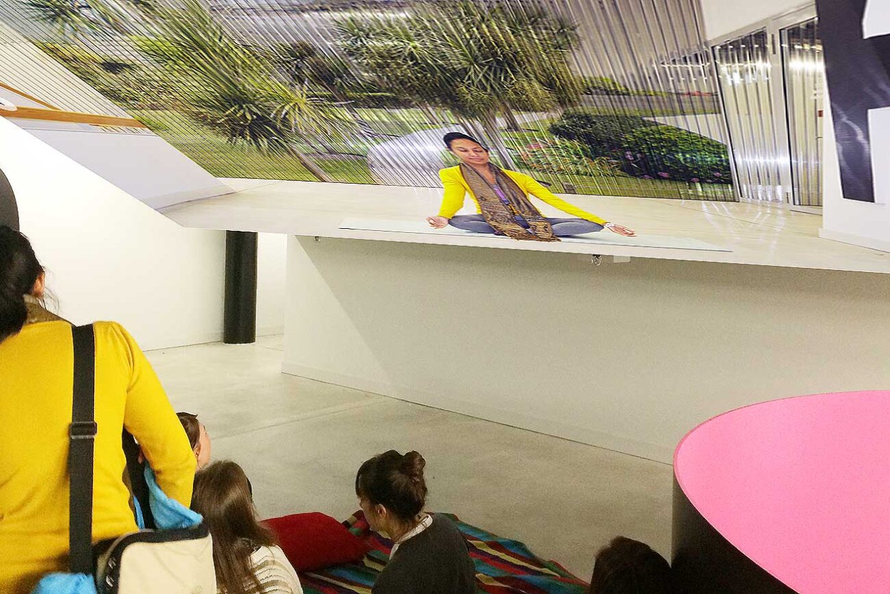 Besucher:innen sitzen im Toni-Areal vor einer grossformatigen Fotoinstallation, die eine meditierende Frau in gelber Jacke in einer parkartigen Landschaft zeigt, und lauschen einer Präsentation des Projekts The Dreamy Island.